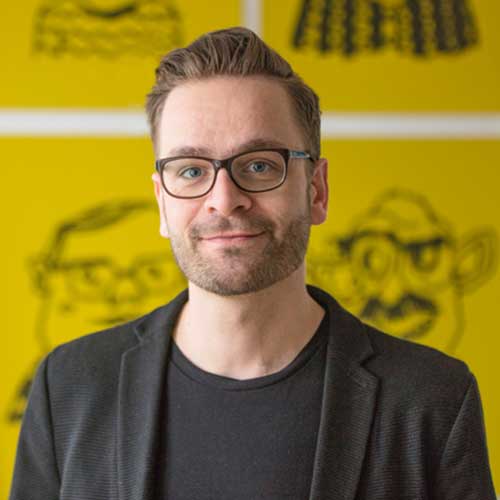 Gregor Strutz vor gelbem Hintergrund. Er trägt eine Brille, kurze dunkelblonde Haare und lächelt.